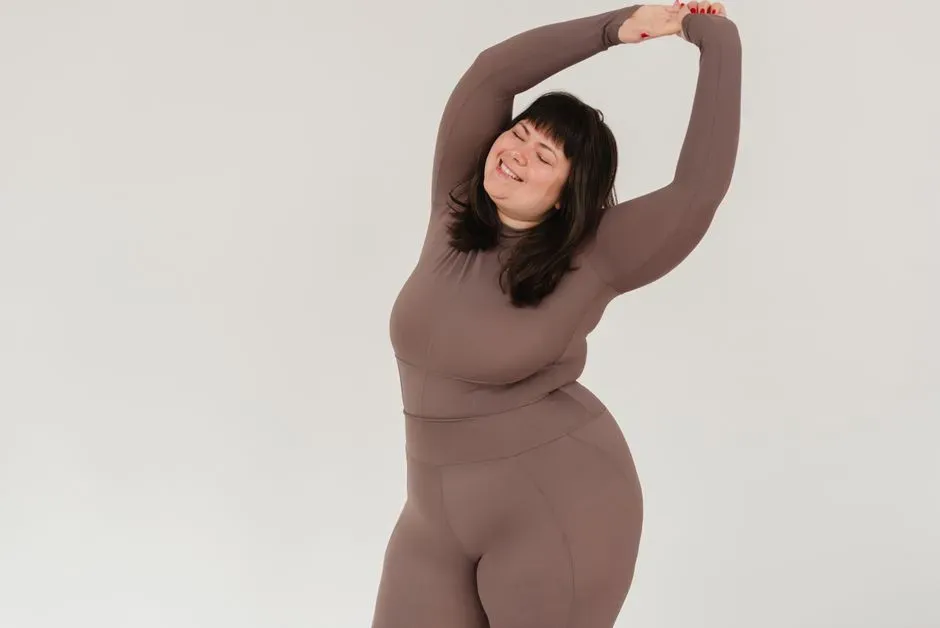 A plus-size woman in a fitted taupe workout set stretching with both arms overhead against a plain white studio background, eyes closed.