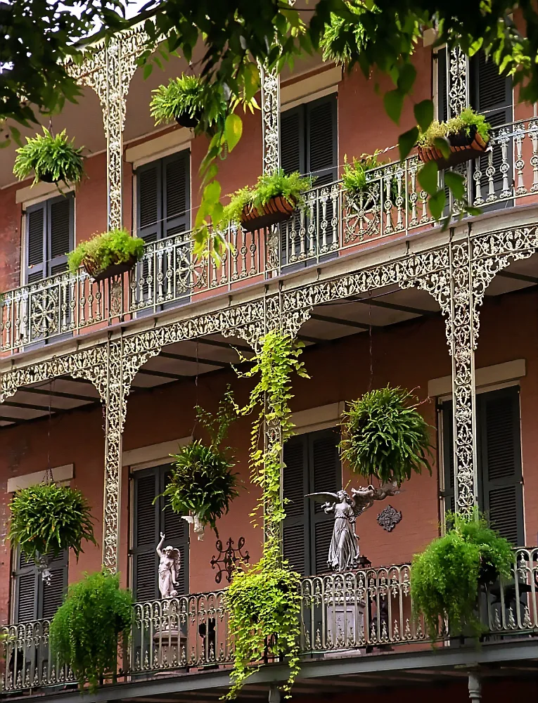 Ornate wrought-iron balconies with hanging fern baskets and two garden statues on a salmon-pink French Quarter building facade in New Orleans, framed by overhanging tree leaves. No people are depicted.