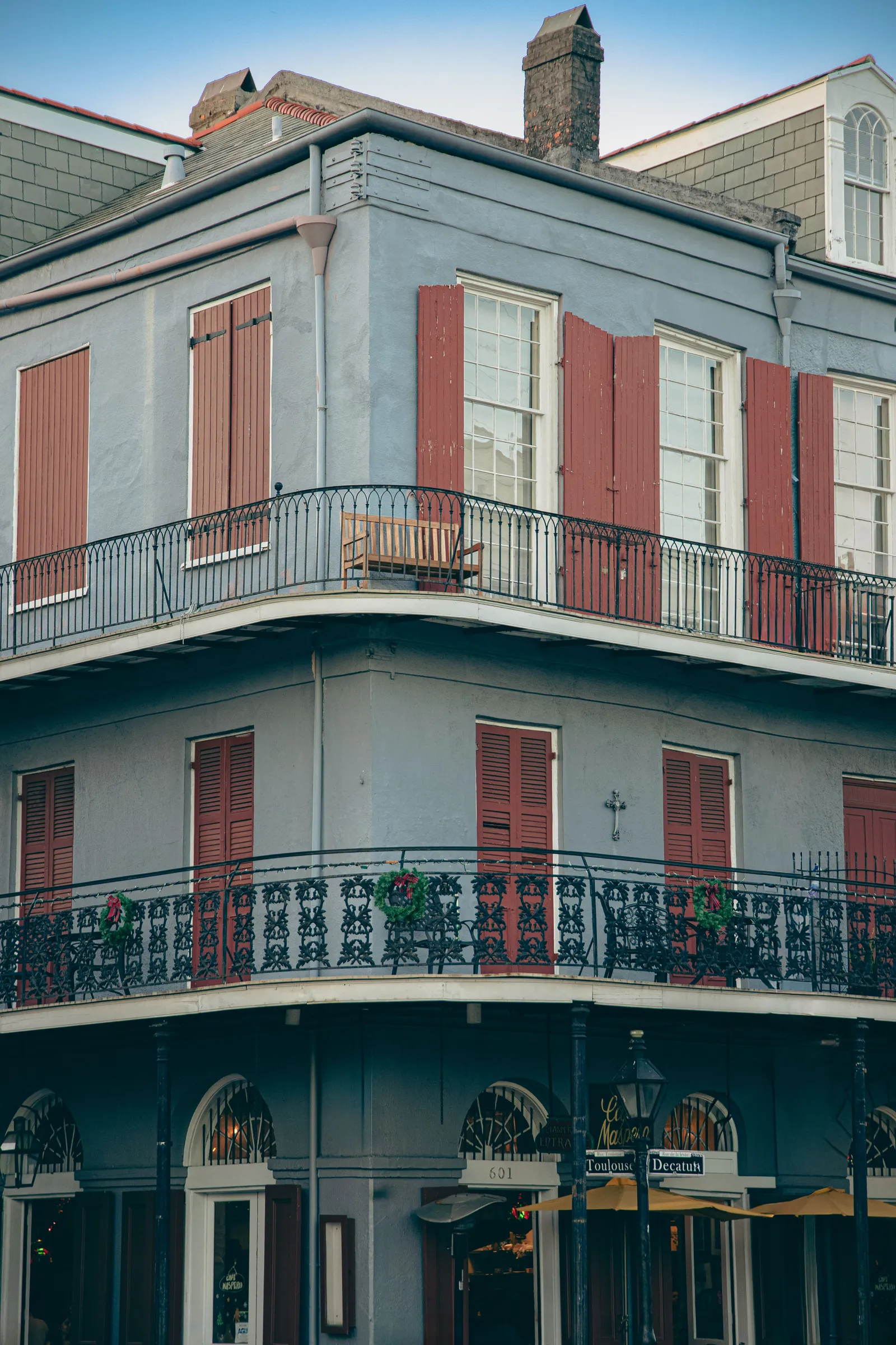 A pale-blue, three-story French Quarter corner building in New Orleans with red shutters, wrought-iron balcony railings hung with holiday wreaths, and ground-floor storefronts at the corner of Toulouse and Decatur streets. No people are depicted.