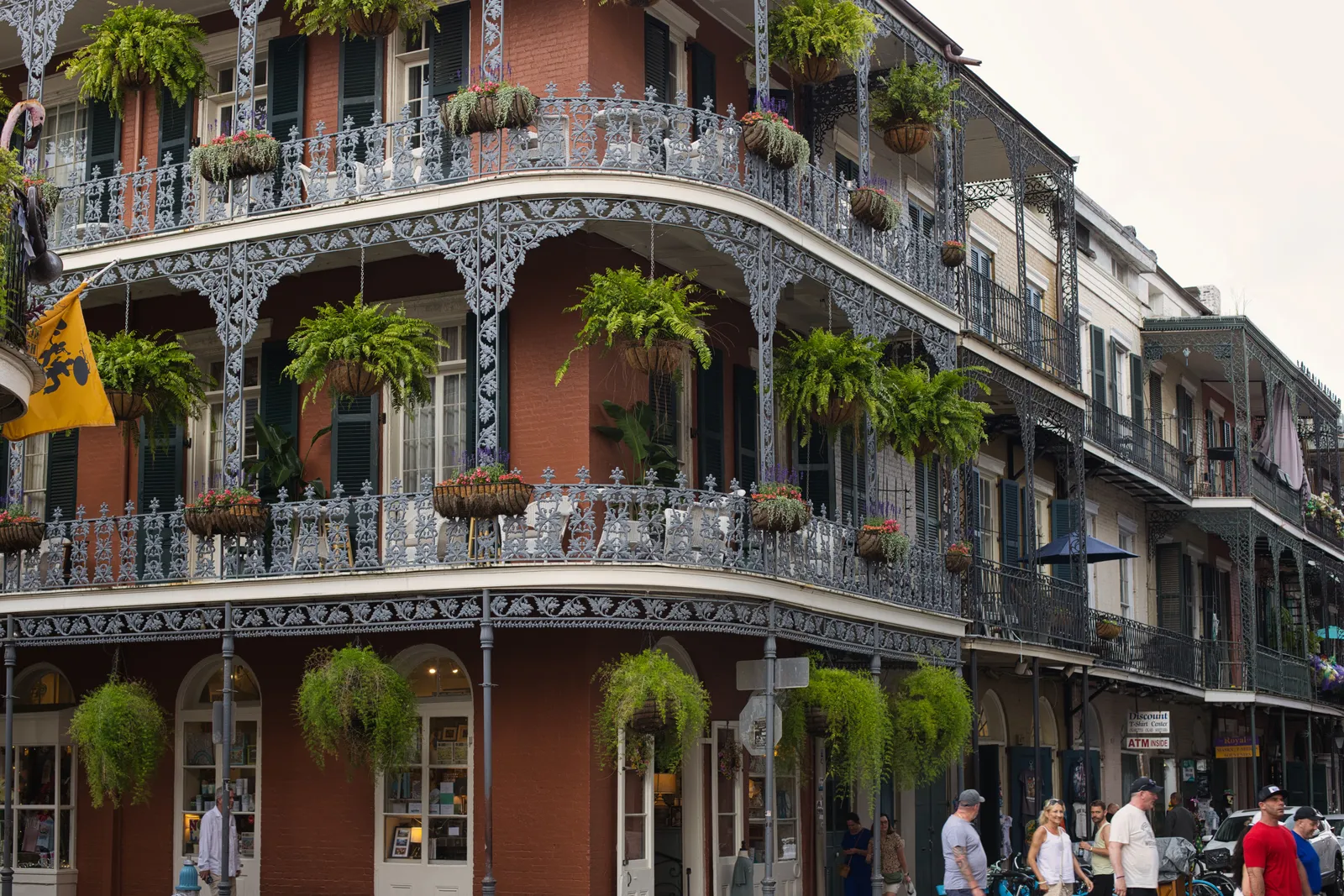 A red-brick French Quarter street corner in New Orleans with three tiers of white wrought-iron balconies hung with fern baskets, storefronts at street level, and a few distant, unidentifiable pedestrians walking the sidewalk below.