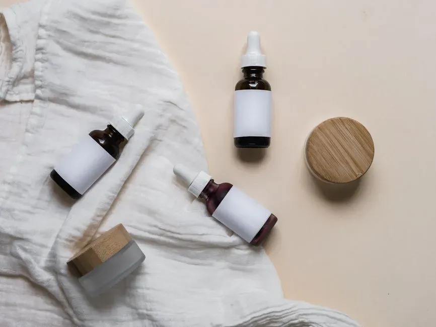 A flat lay of unlabelled cosmetic bottles, droppers and a wooden jar lid arranged on draped white linen.