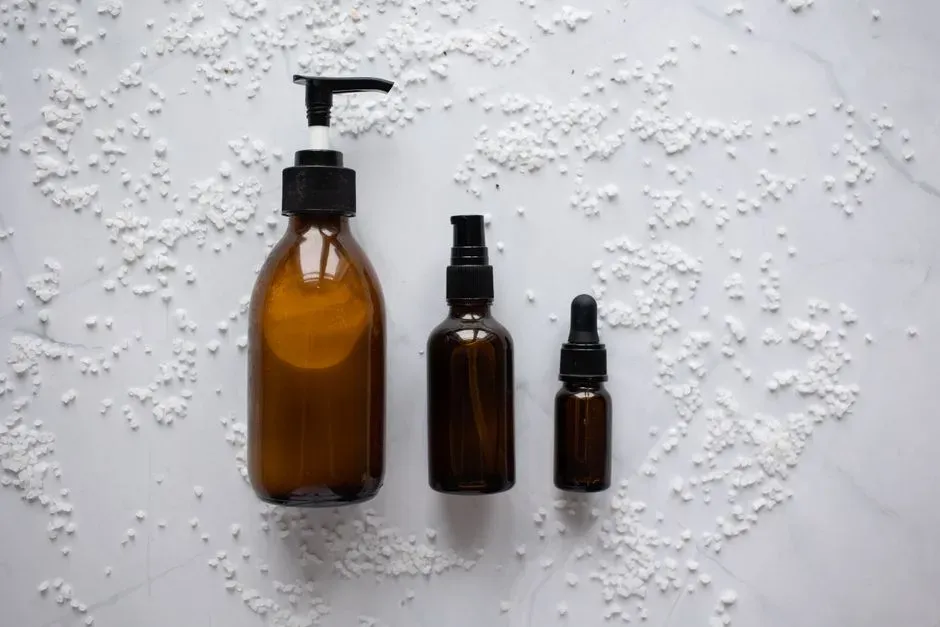 Three amber apothecary-style bottles — a pump, a dropper and a small vial — on a white surface dusted with bath salts.