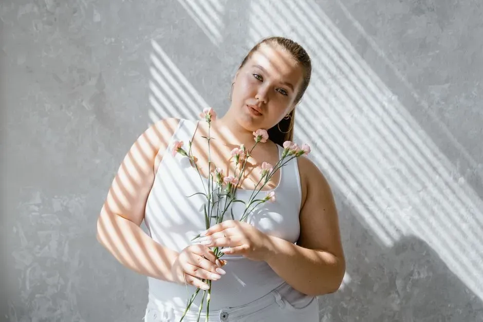 A plus-size woman in a white tank top holding a small bunch of wildflowers, striped shadows falling across her from a window blind.