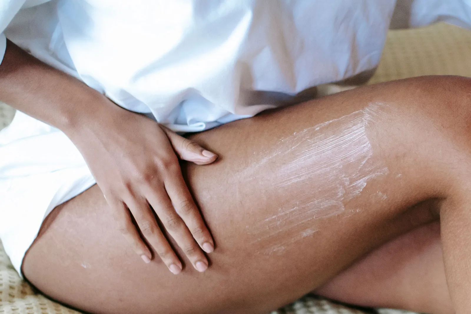 Close-up of a woman's hands smoothing cream along her inner thigh while sitting on a bed — the everyday body-care moment thigh products exist for.