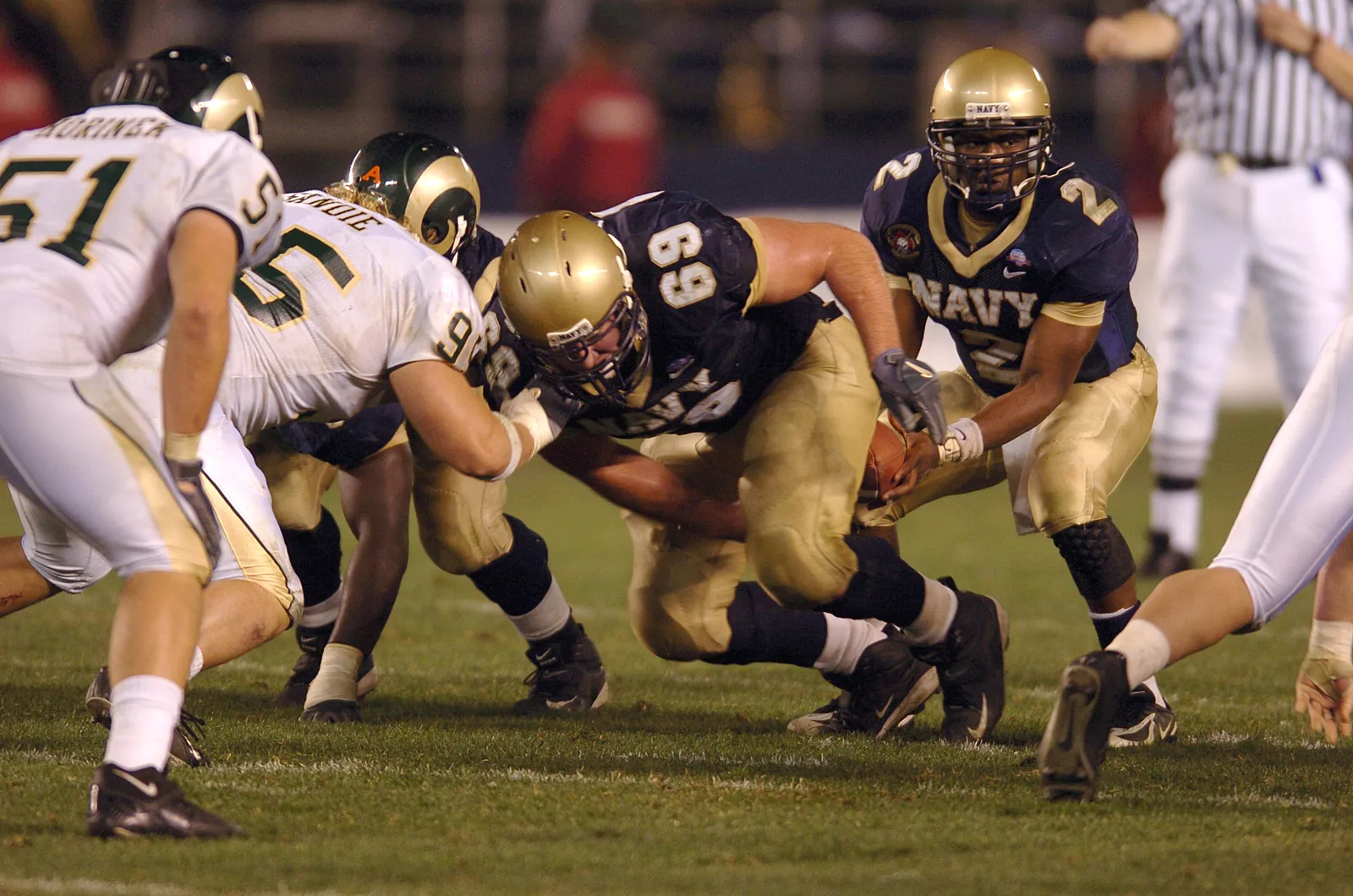 Navy's No. 69 crouches low with the football gripped tight to his body at the line of scrimmage during the 2005 Poinsettia Bowl, as a Colorado State defender grabs on, a Navy teammate closes in, and a referee looks on from behind.