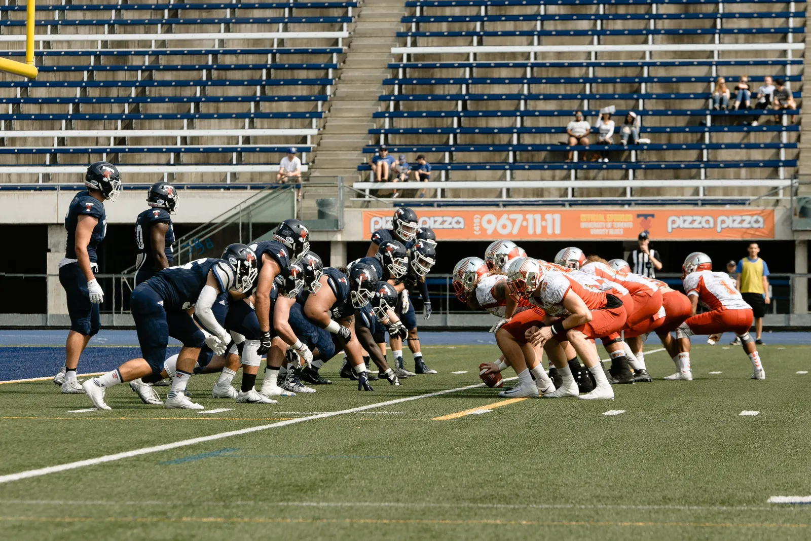 Two football teams, one in navy and one in white and orange, line up at the line of scrimmage in a nearly empty stadium as the center prepares to snap the ball, with a referee and a few spectators visible in the stands behind them.
