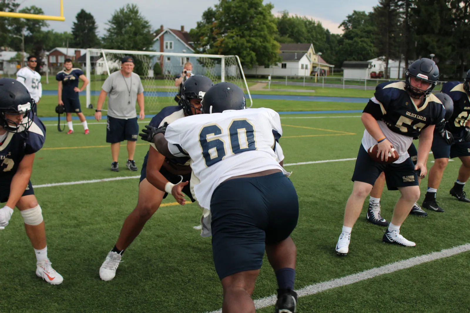 A Clarion Golden Eagles offensive lineman, No. 60, drives into a teammate at a practice session while a quarterback in a Clarion Football jersey holds the ball nearby and a coach watches from the sideline.