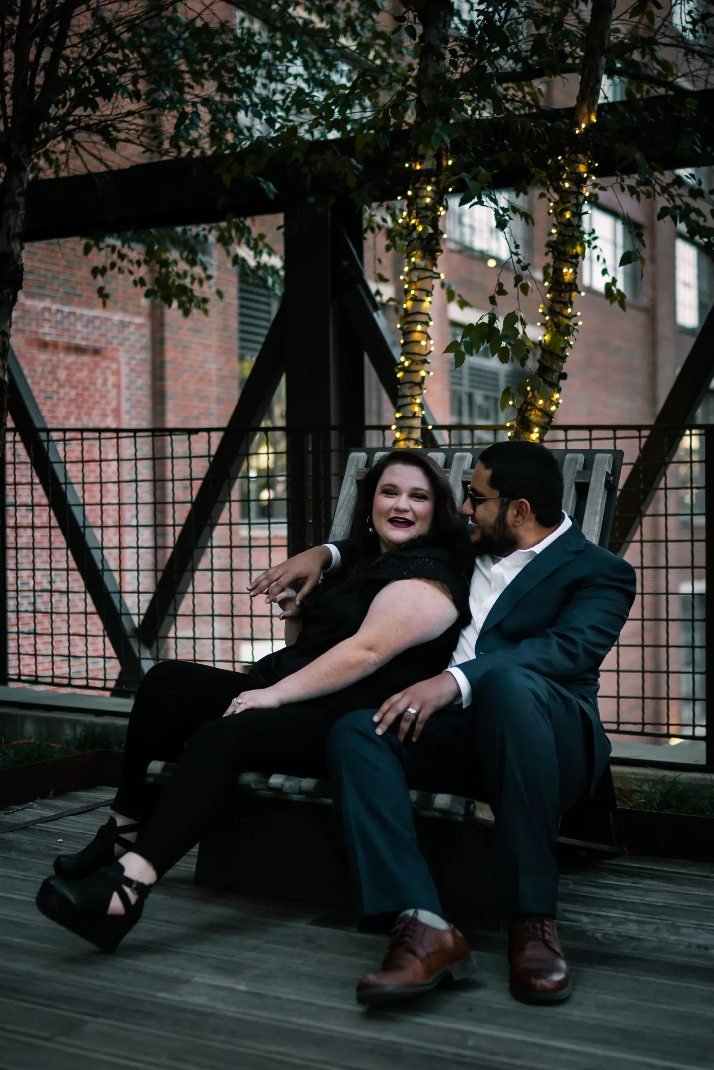 A couple sitting close together on a bench on a string-lit terrace at dusk, laughing - a plus-size woman in black leaning back against a slimmer man in a dark suit, his arm around her.