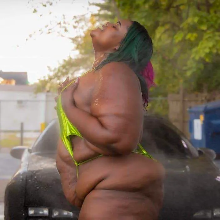 Mzbogotti standing in profile in front of the black car, head tipped back and eyes closed, water spraying in the low sun, the green swimsuit pulled aside and one arm drawn across her chest.
