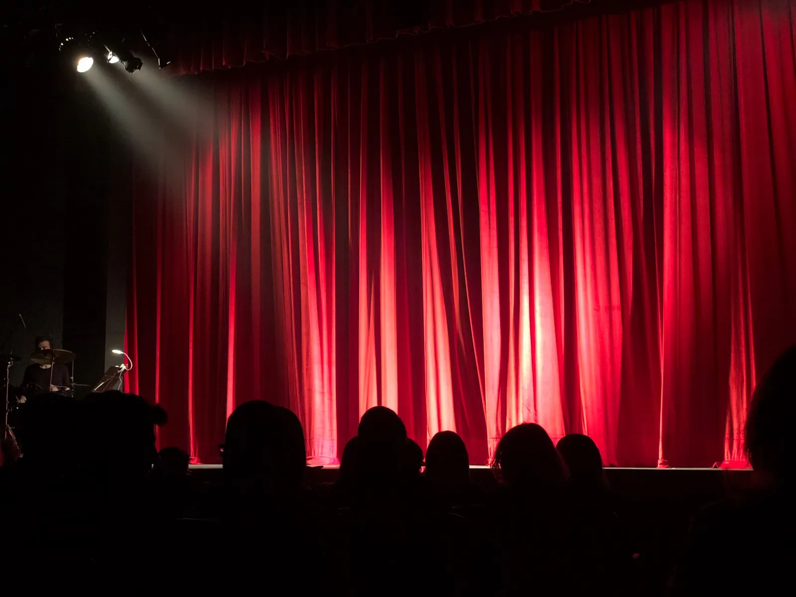 A dimly lit theater stage with red curtains, seen from behind rows of audience members shown only in silhouette, no face identifiable.