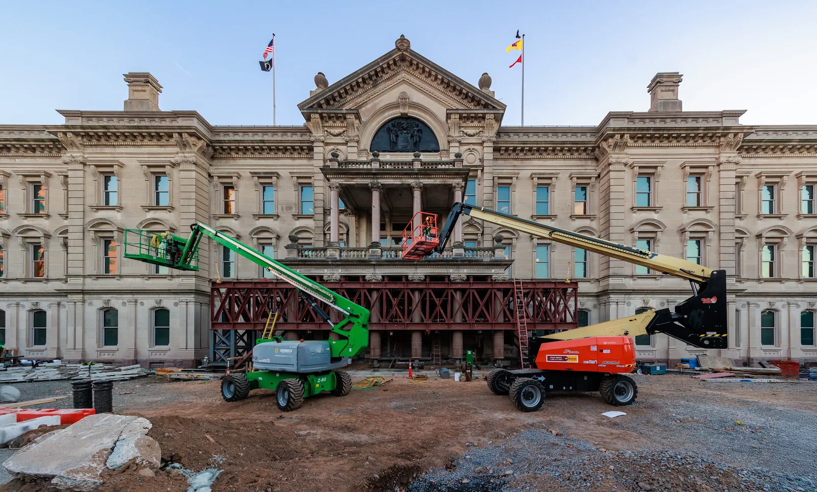 The front of the New Jersey State House mid-renovation, with green and orange cherry pickers parked on churned ground in front of the scaffolded facade and flags flying from the roofline.