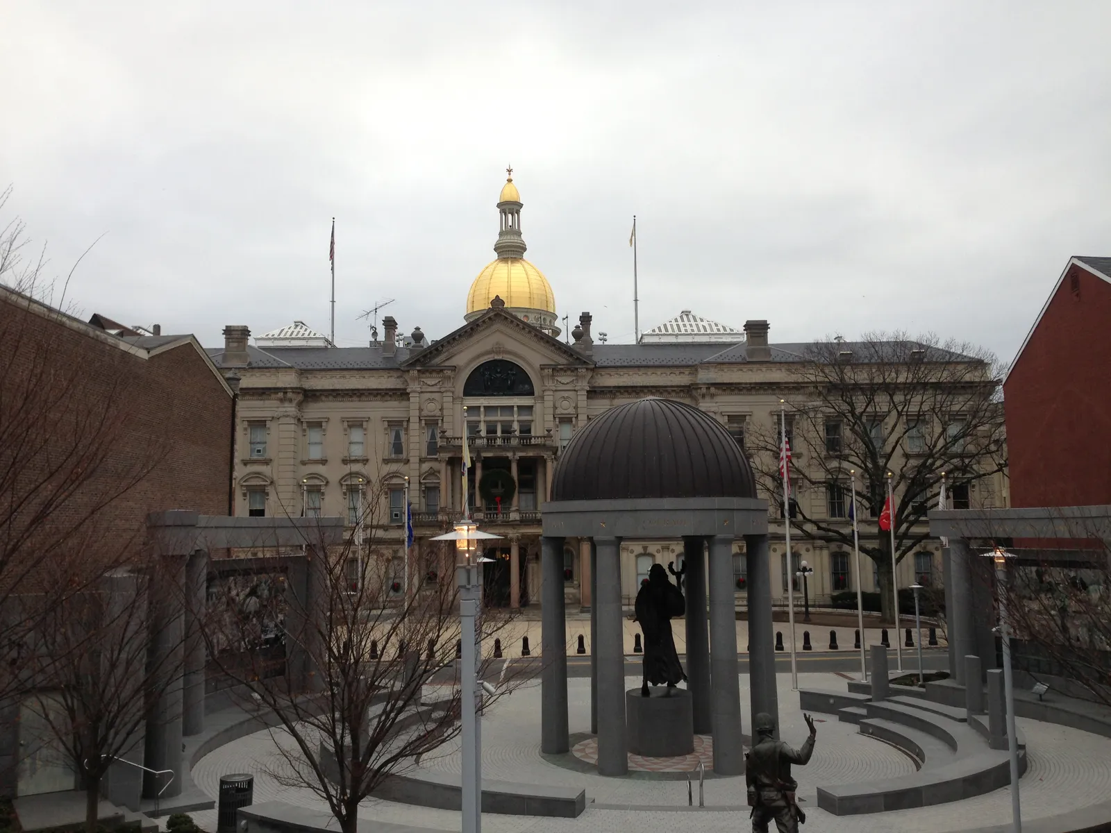 The New Jersey State House in Trenton photographed from across its forecourt on an overcast winter day, its gilded dome rising above the columned entrance, with the domed rotunda of the state World War II Memorial in the foreground.