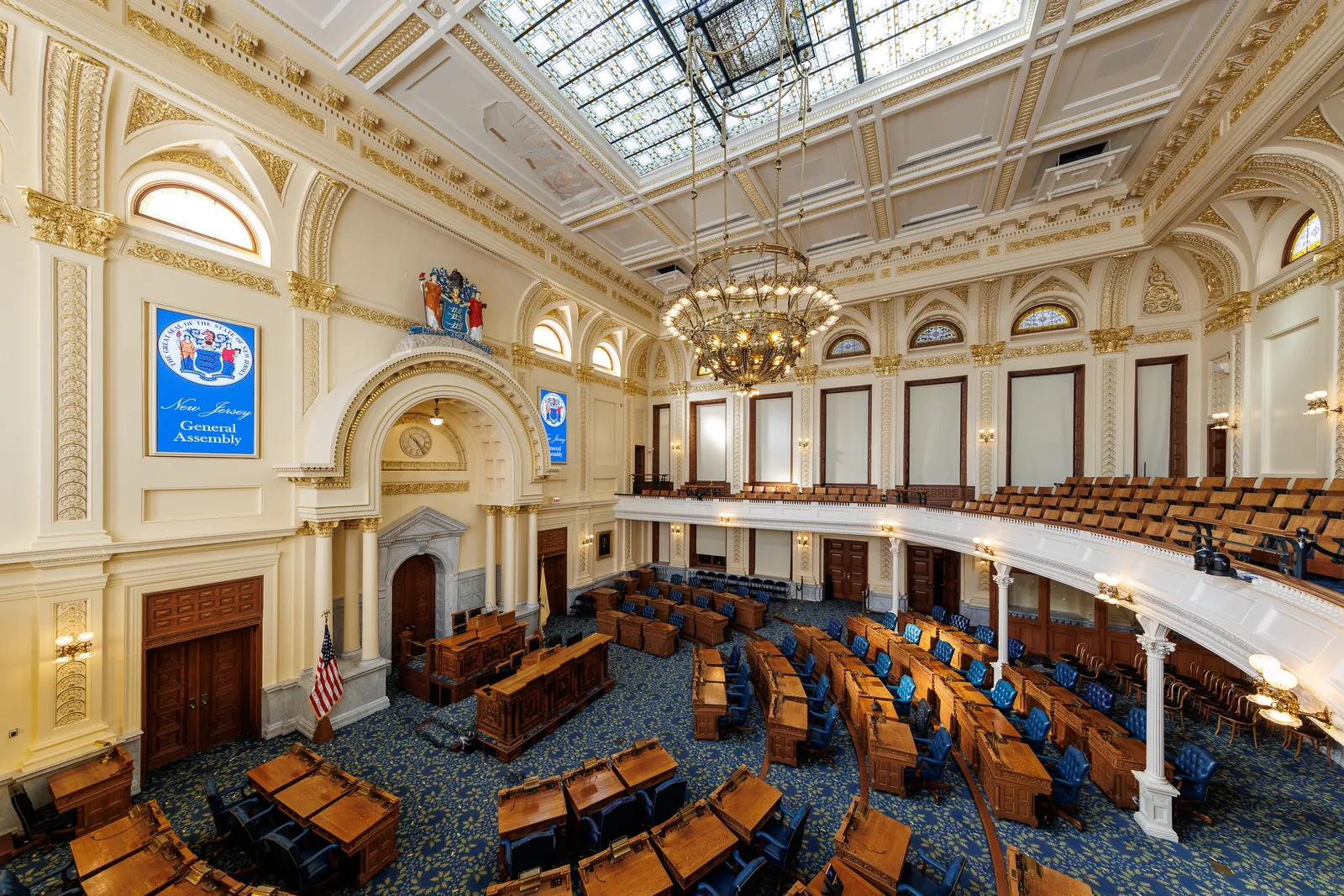 An elevated interior view of the New Jersey General Assembly chamber looking toward the rostrum beneath an arched skylight, with illuminated blue "New Jersey General Assembly" signage on the left wall, a wrought chandelier hanging from the coffered ceiling, and rows of empty wooden legislative desks with blue upholstered chairs and an empty public gallery above.
