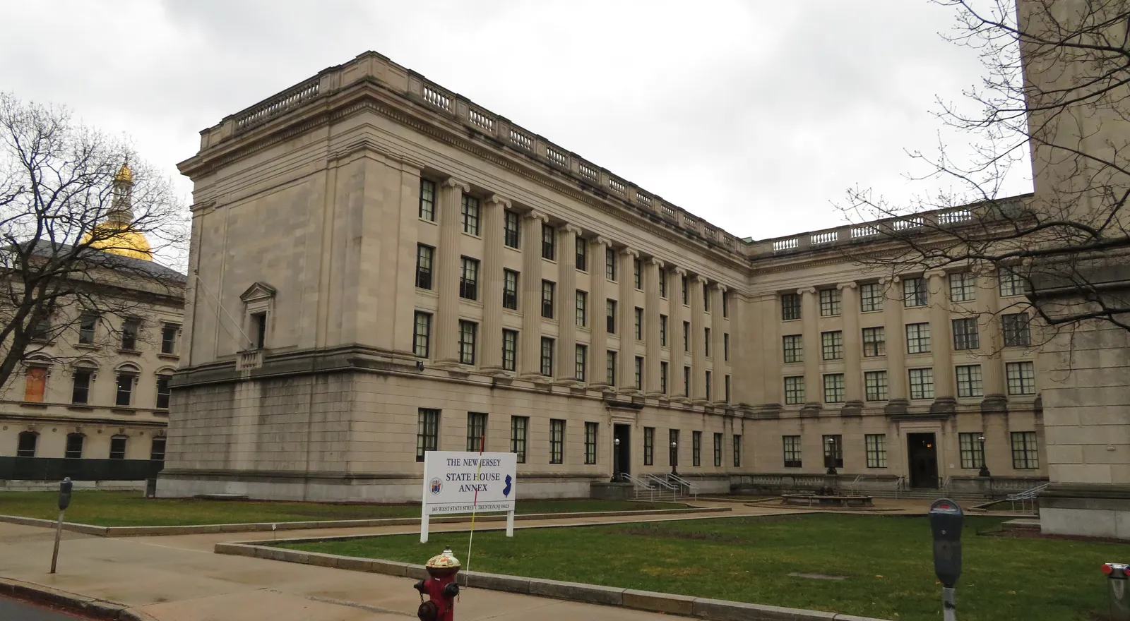 The New Jersey State House Annex in Trenton, a limestone office building with fluted Ionic columns and a balustraded roofline, seen from the street on an overcast day, with a sign reading "The New Jersey State House Annex" on the lawn and the State House's gilded dome visible in the background at left.