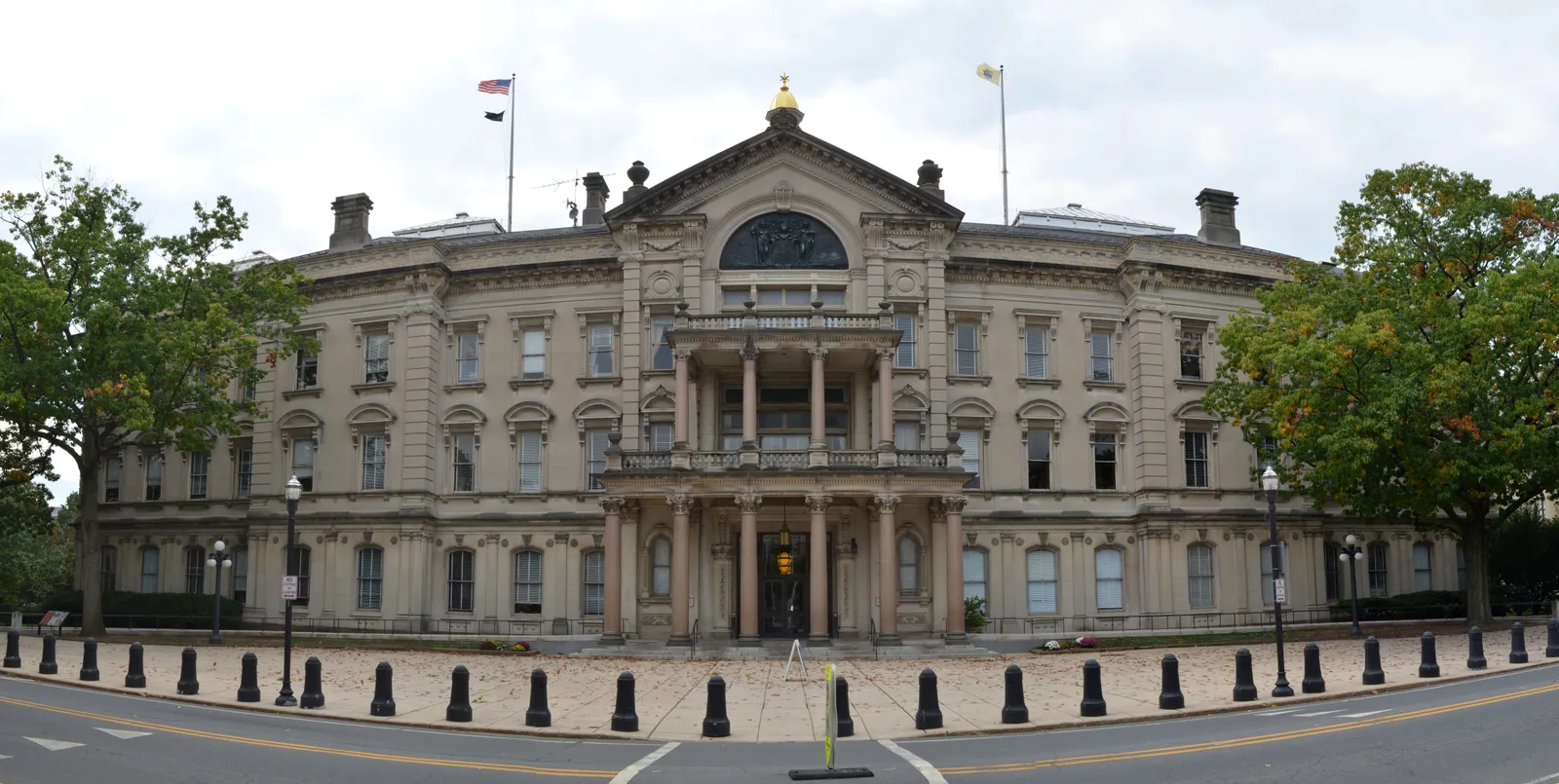 A wide front view of the New Jersey State House in Trenton, its columned portico and gilded dome rising above flanking wings, framed by trees on either side under an overcast sky.