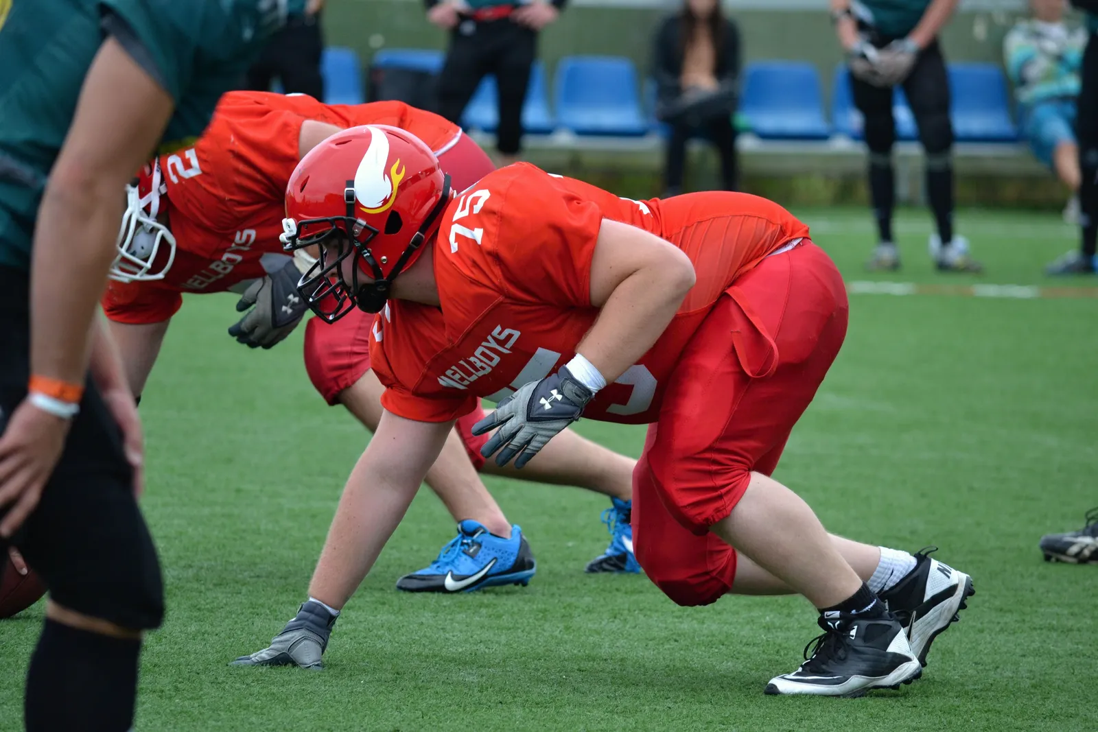 A lineman in a red jersey and helmet crouches into a three-point stance at the line of scrimmage, with a teammate beside him and empty blue stadium seating in the background.