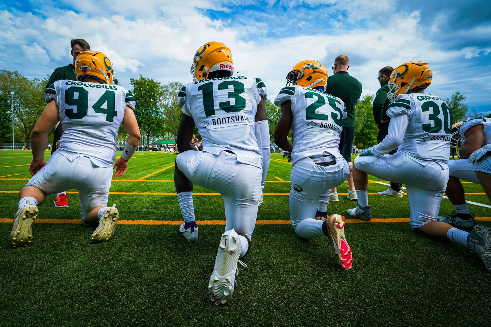 Four football players in white 'Crocodiles' jerseys numbered 94, 13, 25 and 30 kneel in a row on one knee, seen from behind, with teammates, coaches and a cloudy sky beyond the field.