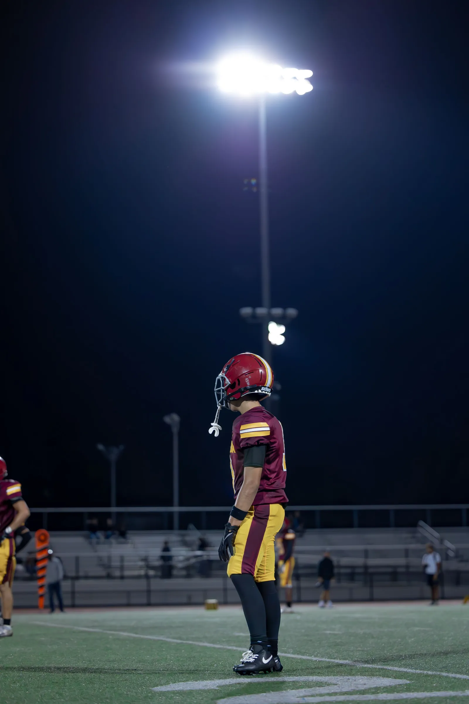 An American football player in a red helmet and maroon-and-gold uniform stands on the field under stadium lights at night, with teammates and sparse bleacher seating visible behind him. No NFL team markings are visible.