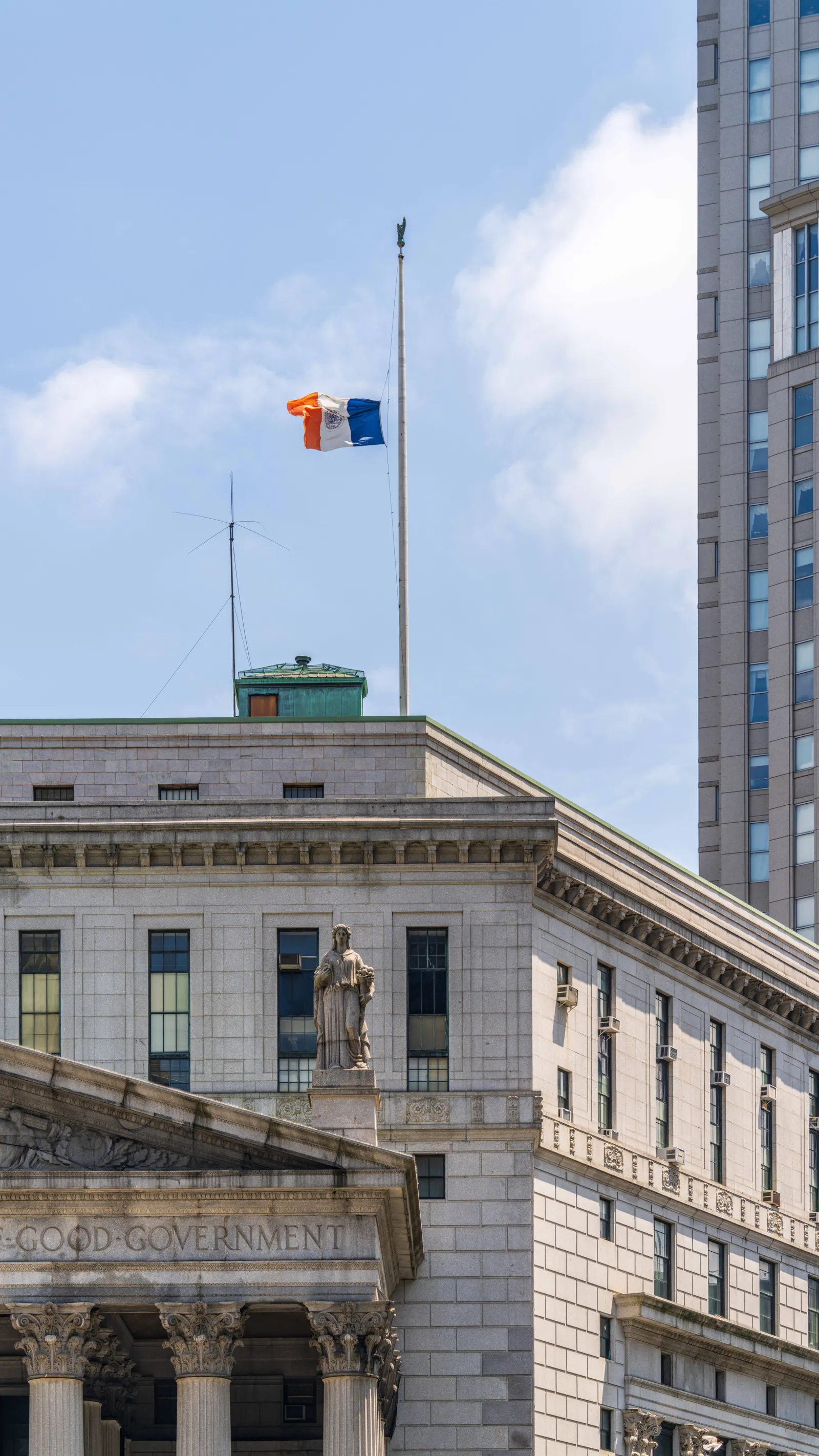 A neoclassical courthouse in New York City, its columned stone facade seen under a bright blue summer sky with a New York City flag flying in front of it.