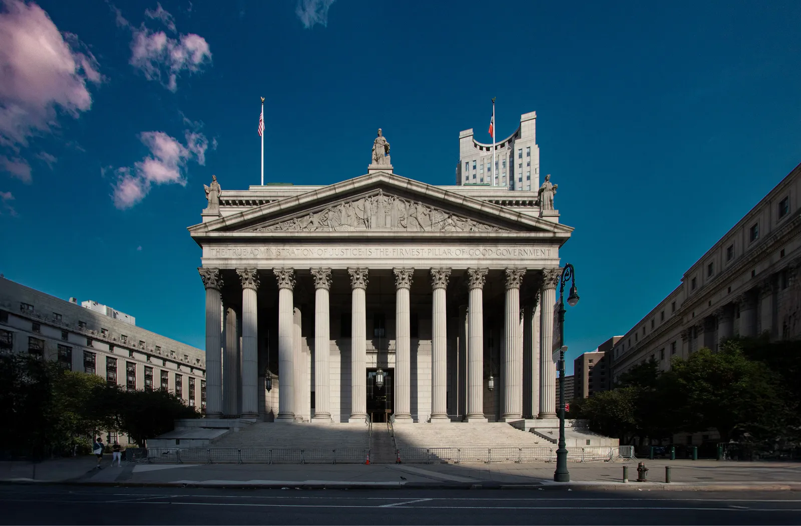 The exterior of a New York Supreme Court building, its columned stone facade photographed head-on beneath a clear blue sky.