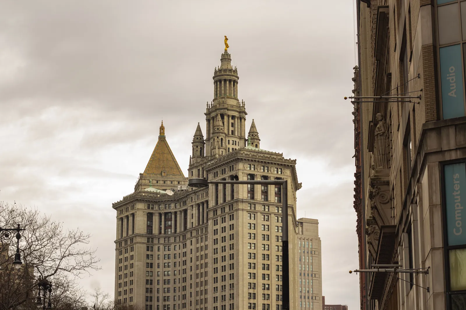 The Manhattan Municipal Building, a tall neoclassical government tower with a colonnaded base and a tiered crown, photographed from street level under a cloudy sky.
