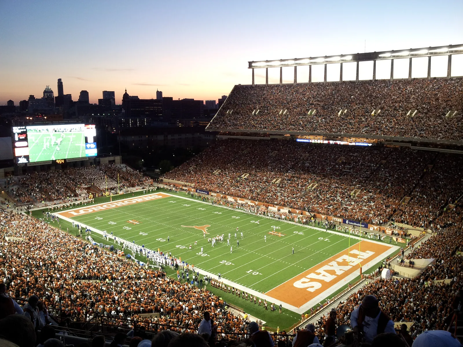 The interior bowl of Darrell K Royal-Texas Memorial Stadium at dusk during a football game, stands packed with fans, the field and video board visible below, and the Austin skyline silhouetted in the background.