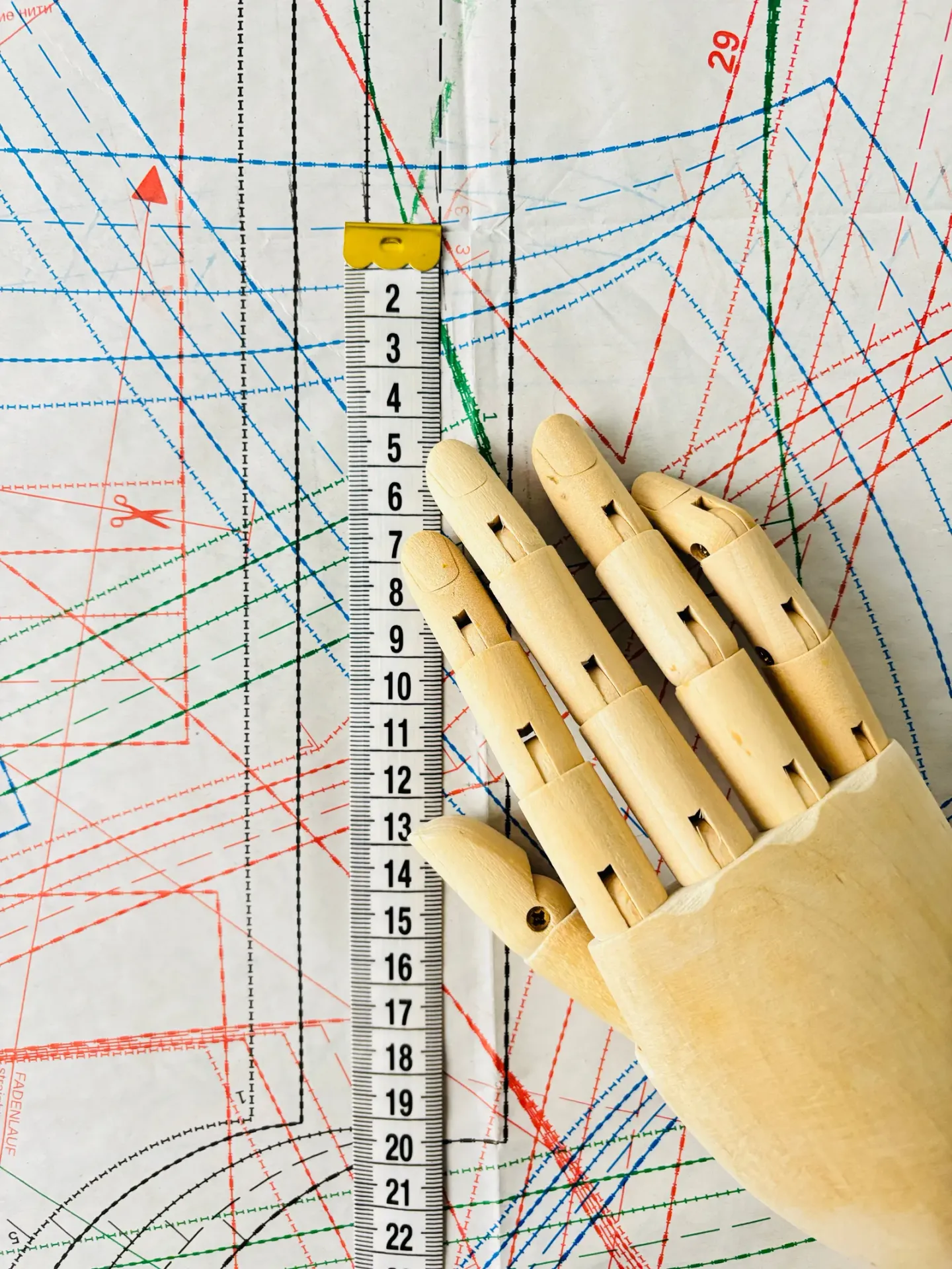 A pattern-drafting sheet covered in overlaid red, blue, green and black grading lines, with a graded measuring scale down the middle and a wooden artist mannequin hand resting on top.