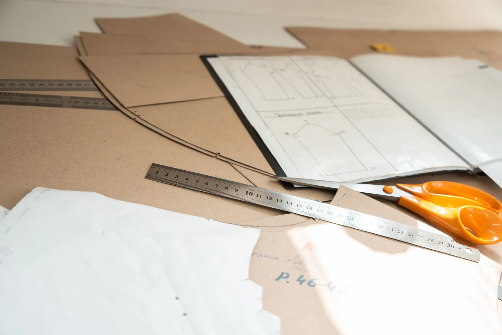 Brown paper pattern pieces marked with drafting lines spread across a work table with a steel ruler, a pencil and orange-handled shears.