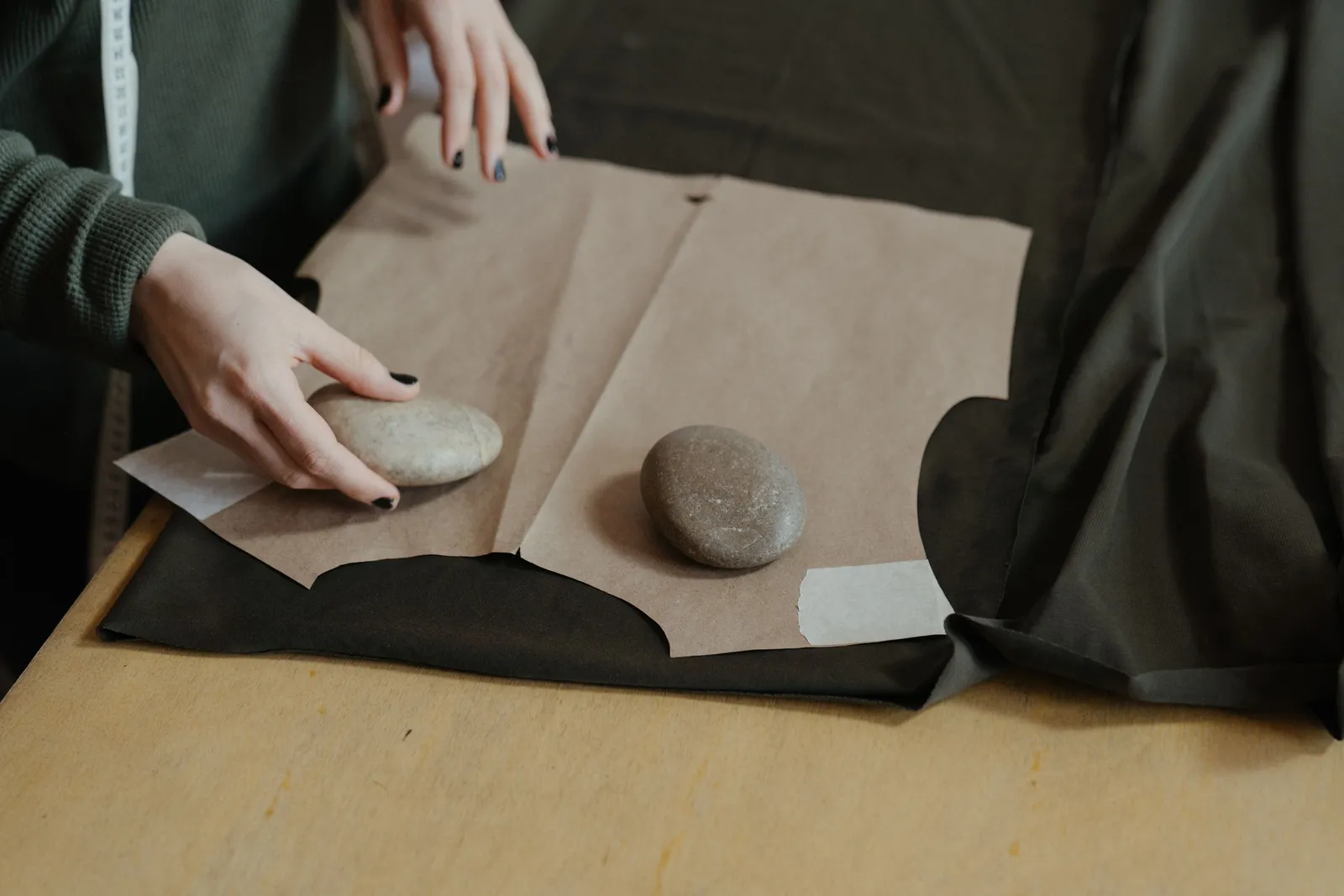 A dressmaker laying a curved brown paper pattern piece onto dark fabric, holding it flat with smooth stone pattern weights.