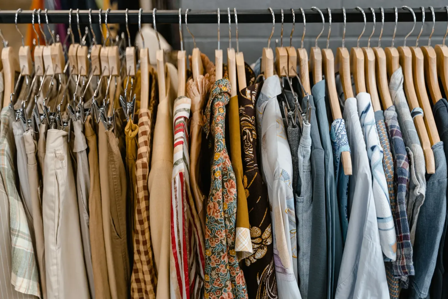 A long rail of clothing on wooden hangers — neutral trousers, patterned shirts, denim and knitwear packed tightly together — running the width of the frame in a store.