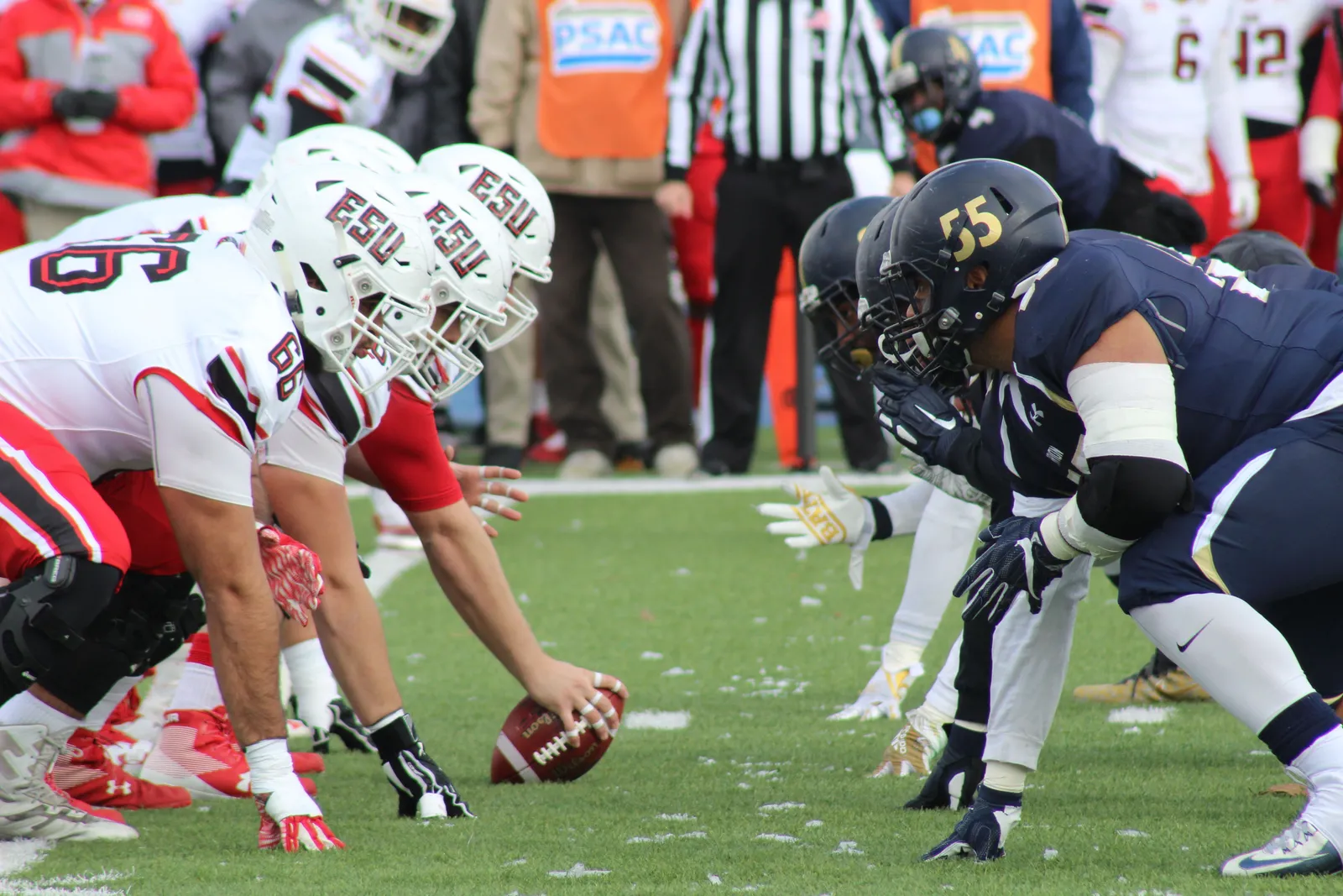 A center in a navy jersey crouched over the football with his hand on the laces, the opposing line set a yard away, in the moment before the snap.