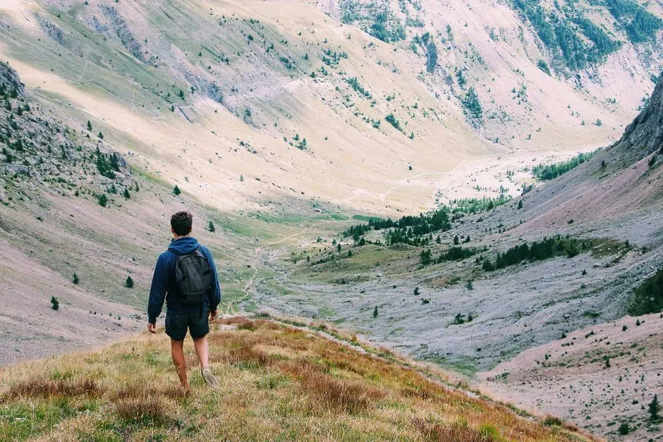A hiker with a backpack walking down a grassy ridge into a broad glacial valley between scree-covered mountains.