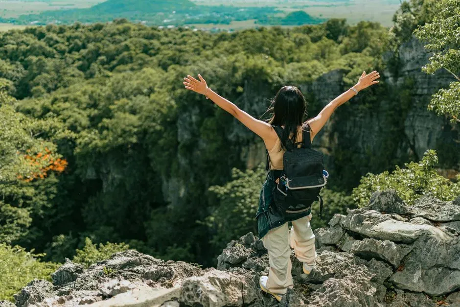 A woman with a backpack standing on a rocky overlook with both arms raised wide toward a forested gorge below.