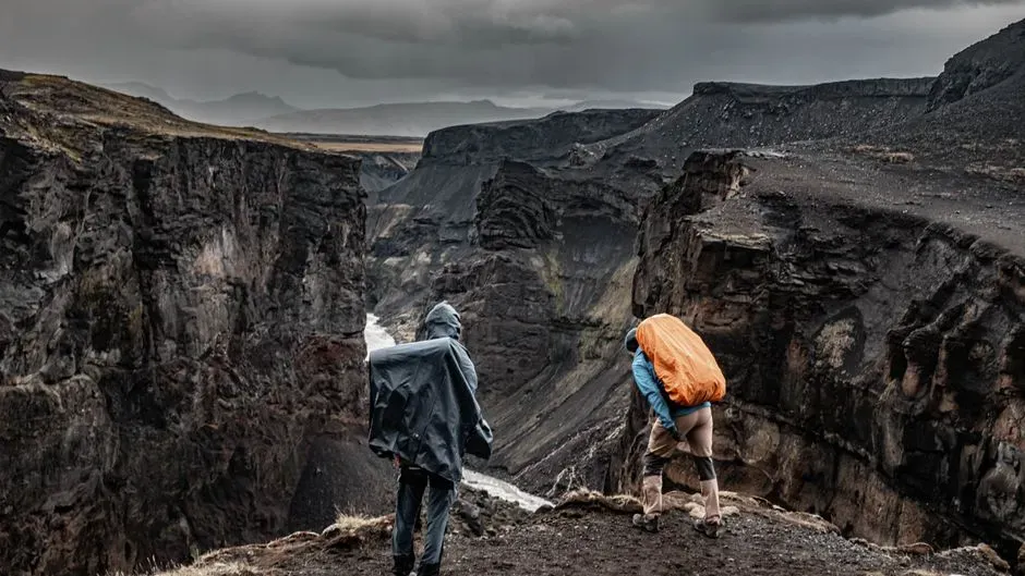 Two hikers in rain shells looking out over a dark, sheer river canyon under an overcast sky.