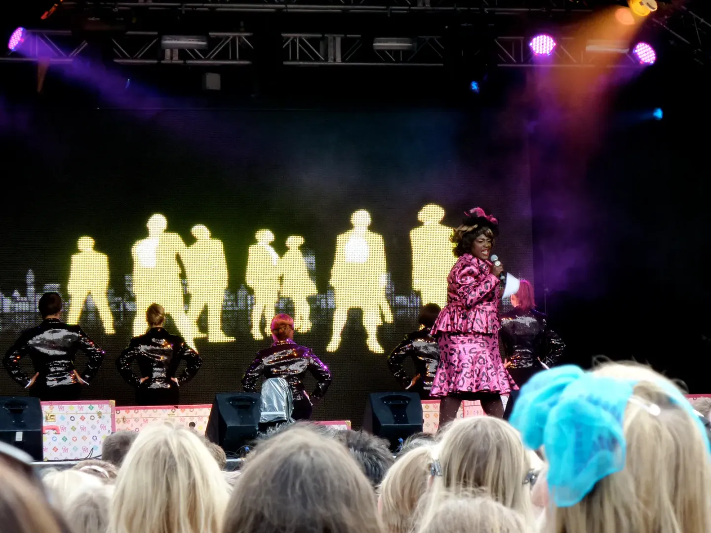 Sharon D Clarke performing on an outdoor stage, microphone in hand, in a pink leopard-print coat and matching hat against a bank of stage lights.