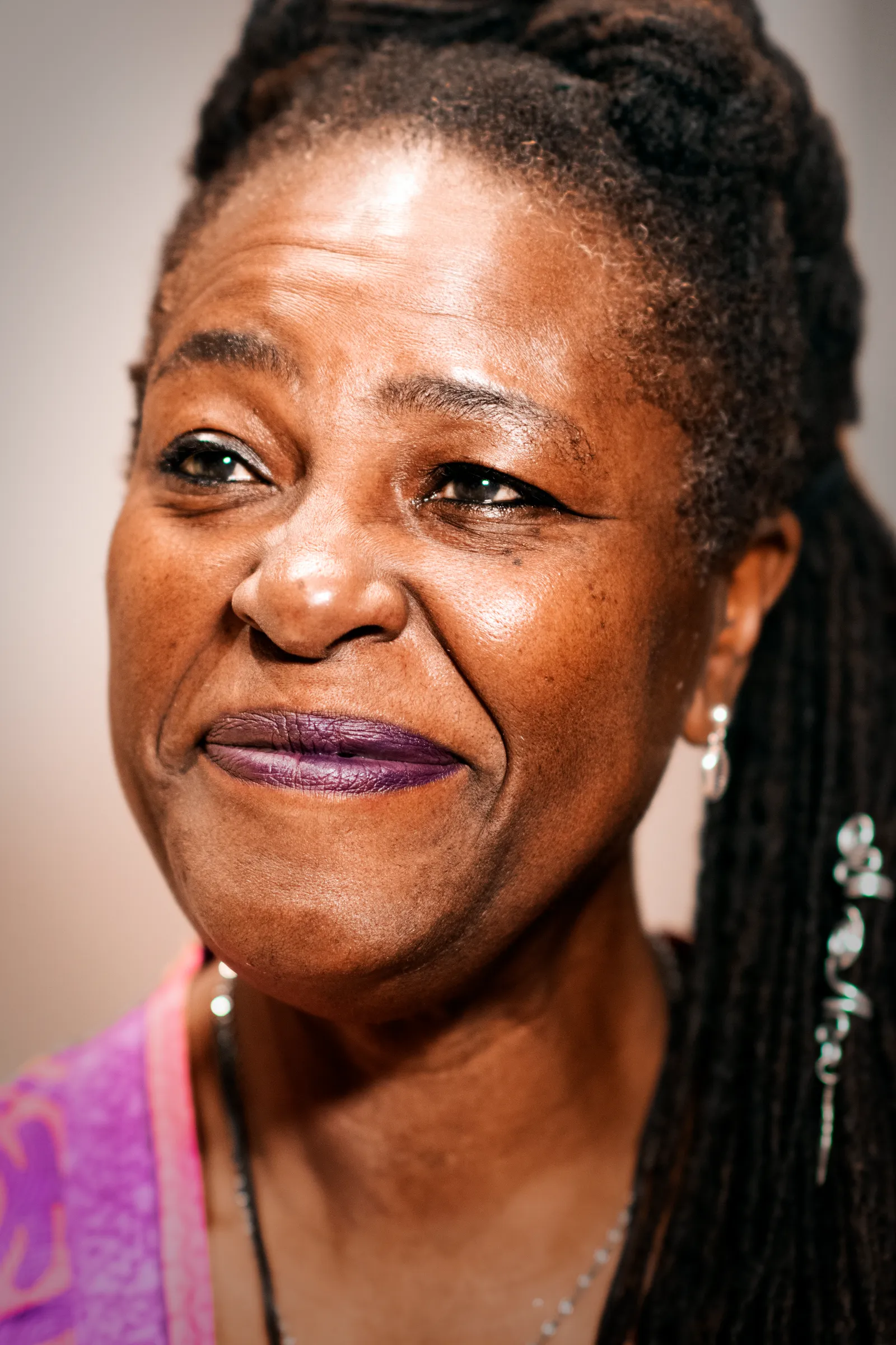 A portrait of Sharon D Clarke smiling to camera, her locs pulled back, wearing drop earrings and a bright pink patterned top against a pale background.