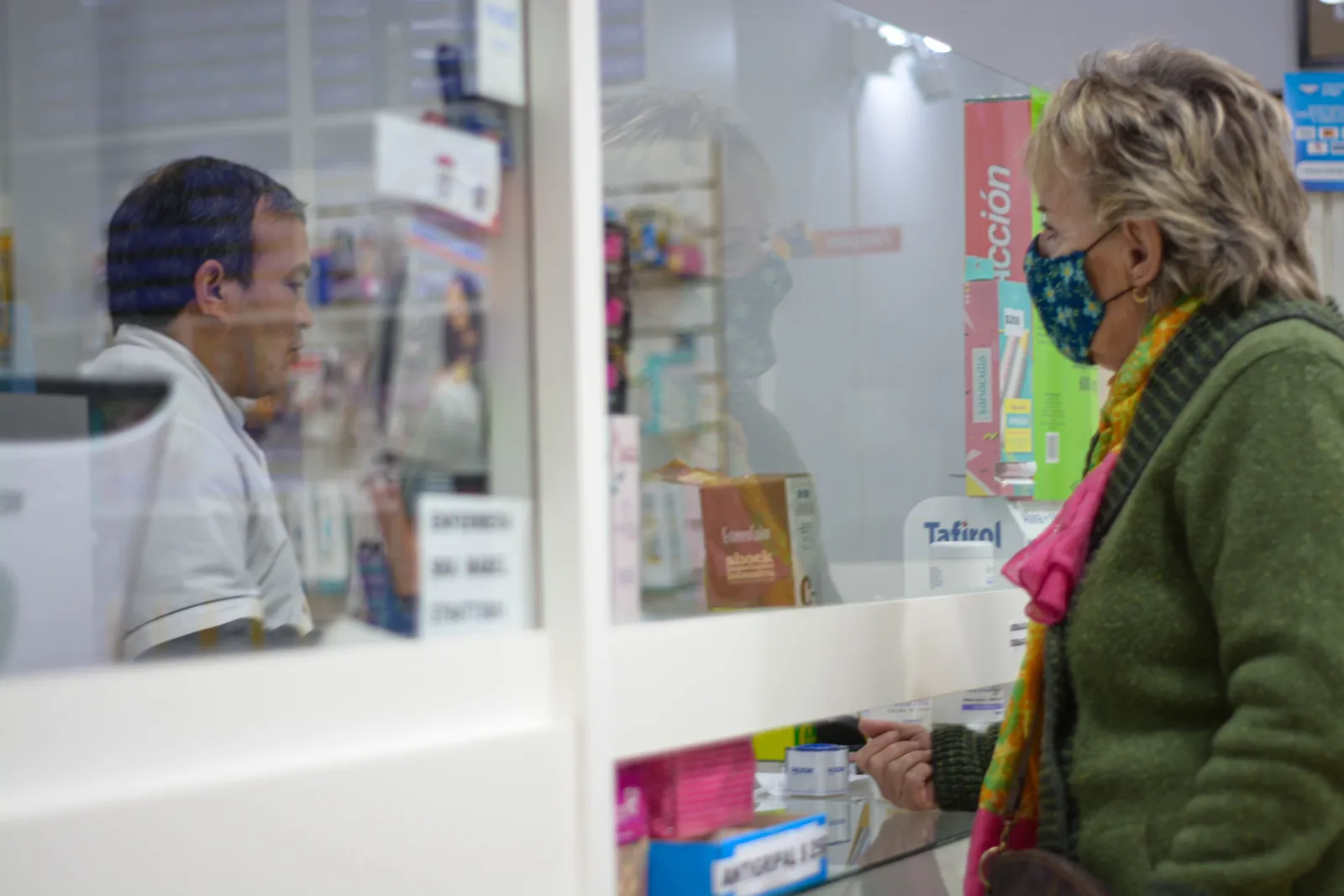 A masked customer speaks with a pharmacist across a glass partition at a pharmacy counter, medication boxes visible on the shelves behind the glass.