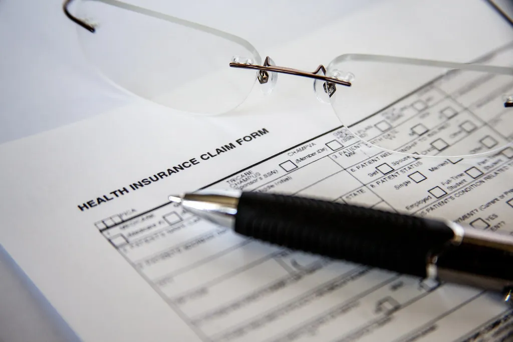 A close-up of a health insurance claim form with a pen and a pair of eyeglasses resting on top of it.