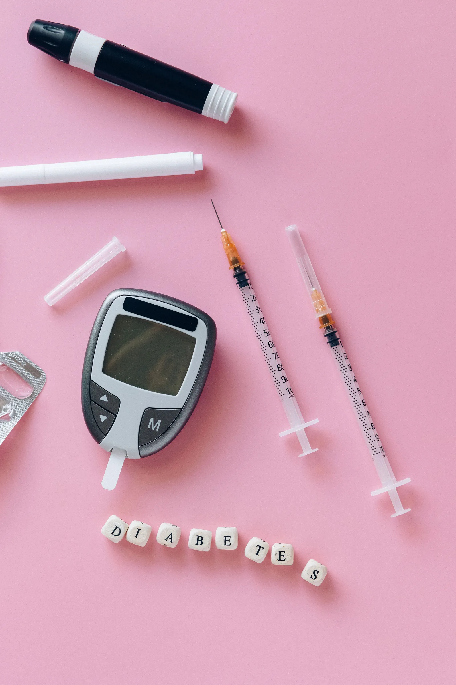 A close-up of diabetes monitoring and injection supplies, including syringes, arranged beside small wooden blocks spelling out "diabetes" on a pink background, with no person or body in frame.