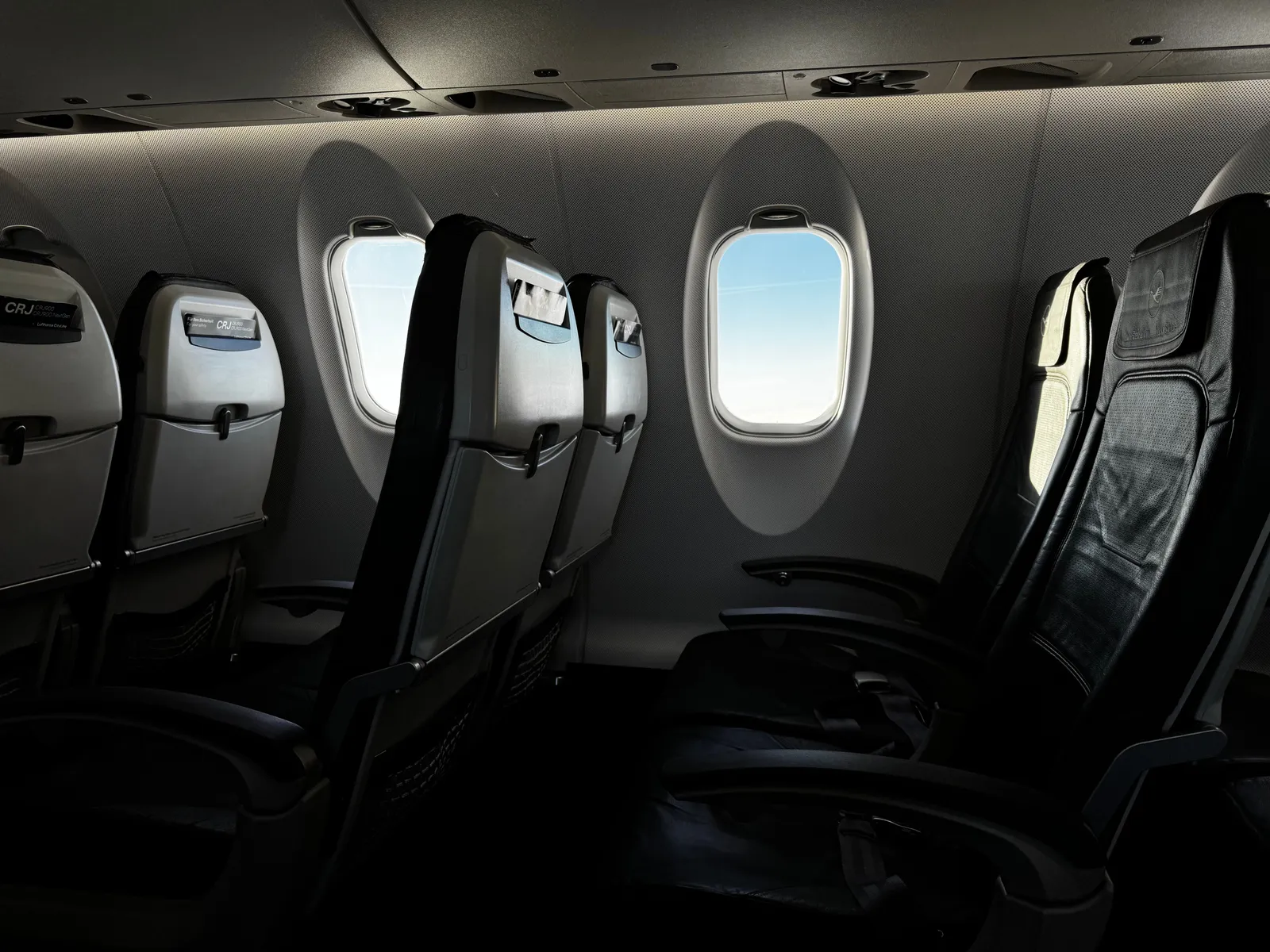 Rows of empty dark aircraft seats beside oval cabin windows, photographed across the row so the narrow gap between armrests is visible.