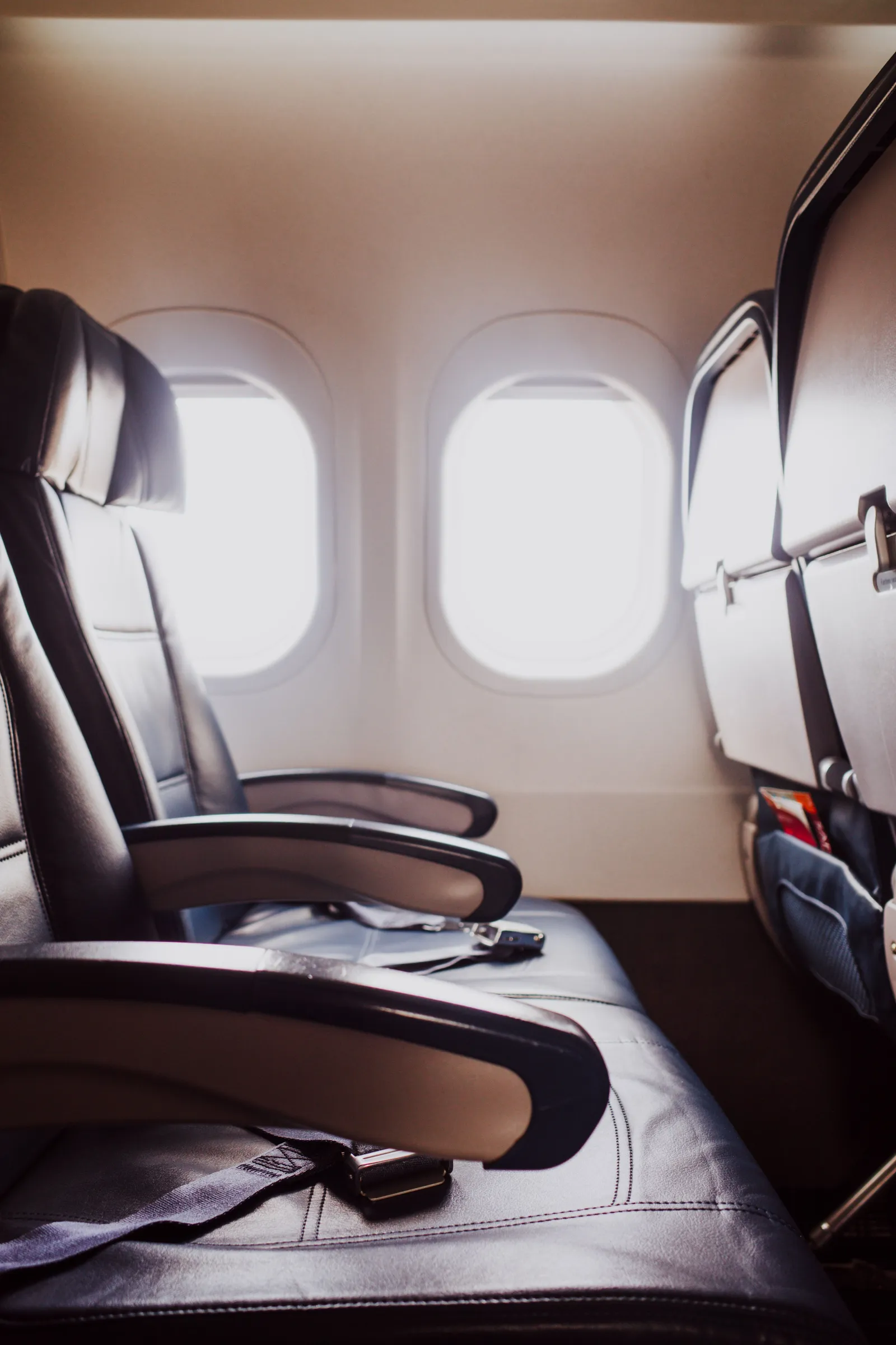 An empty window seat and middle seat on an aircraft, seatbelts laid across the cushions, bright daylight coming through the two windows.