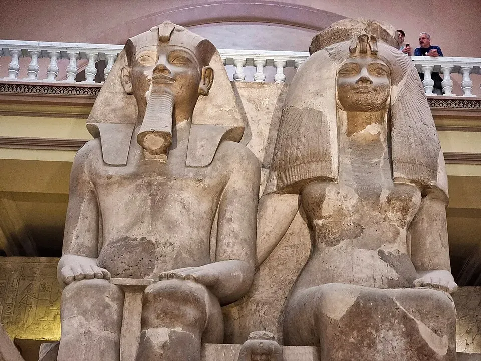 The colossal limestone statue of Amenhotep III and Queen Tiye seated side by side in a museum hall, photographed from below. Both sit upright with hands on their knees, the king in a striped nemes headdress and the queen in a heavy tripartite wig, carved at the same monumental scale.