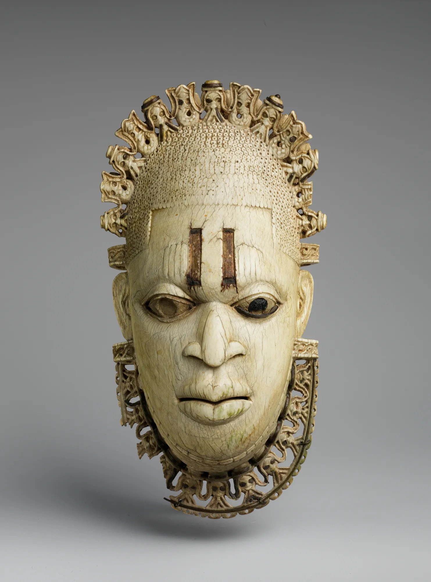 The ivory pendant mask of Ìyọ́bà Idià of Benin, carved as a serene female face with vertical scarification marks above the eyes and inlaid metal pupils, crowned and collared by carved rows of small bearded heads.