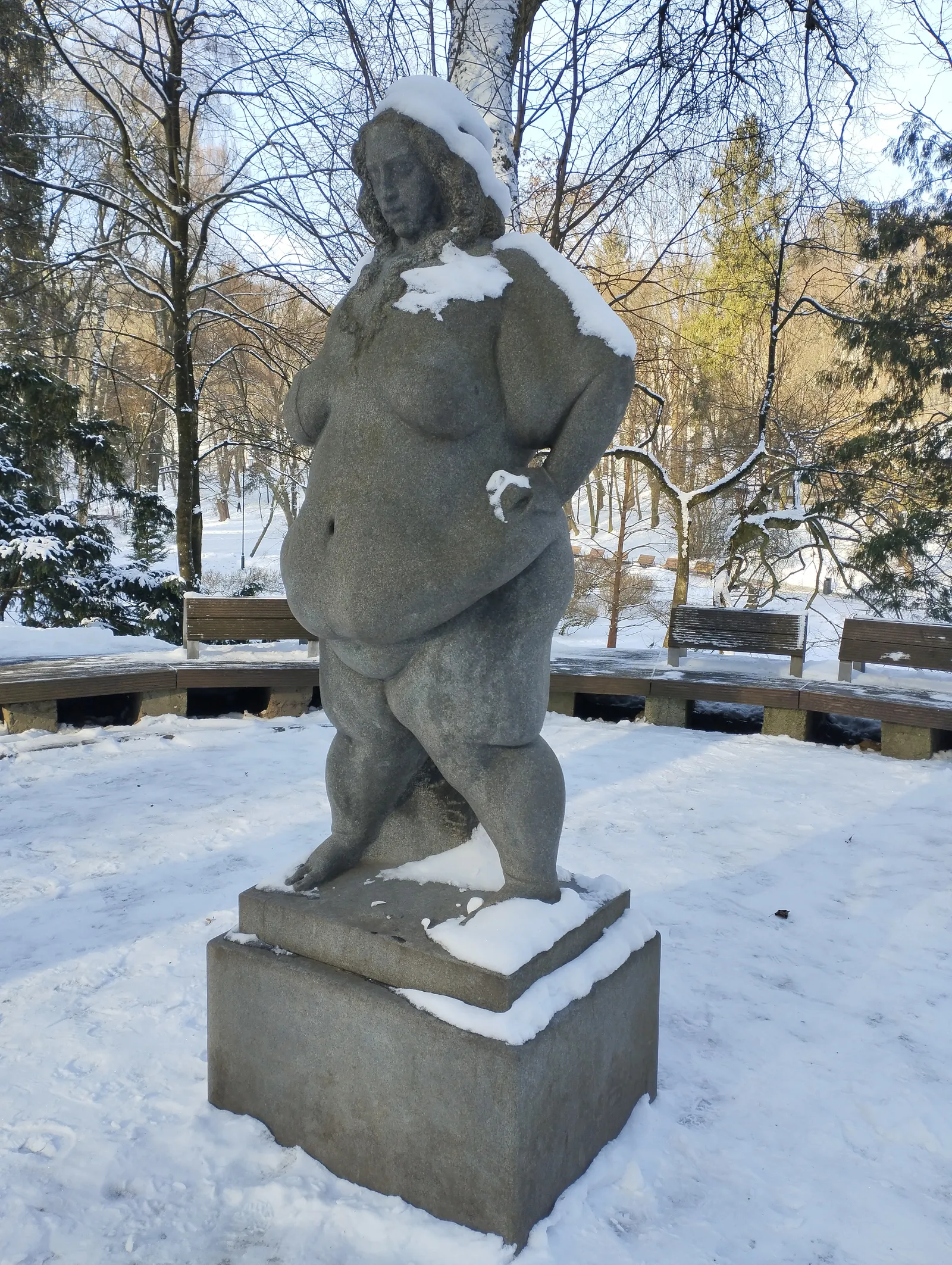 A three-quarter view of the full-length granite statue 'Confident' on its pedestal in Stryi Park, Lviv: a nude, full-figured woman standing with one hand on her hip, her carved hair falling past her shoulders and dusted with snow, bare winter trees and a curved row of snow-covered park benches behind her.