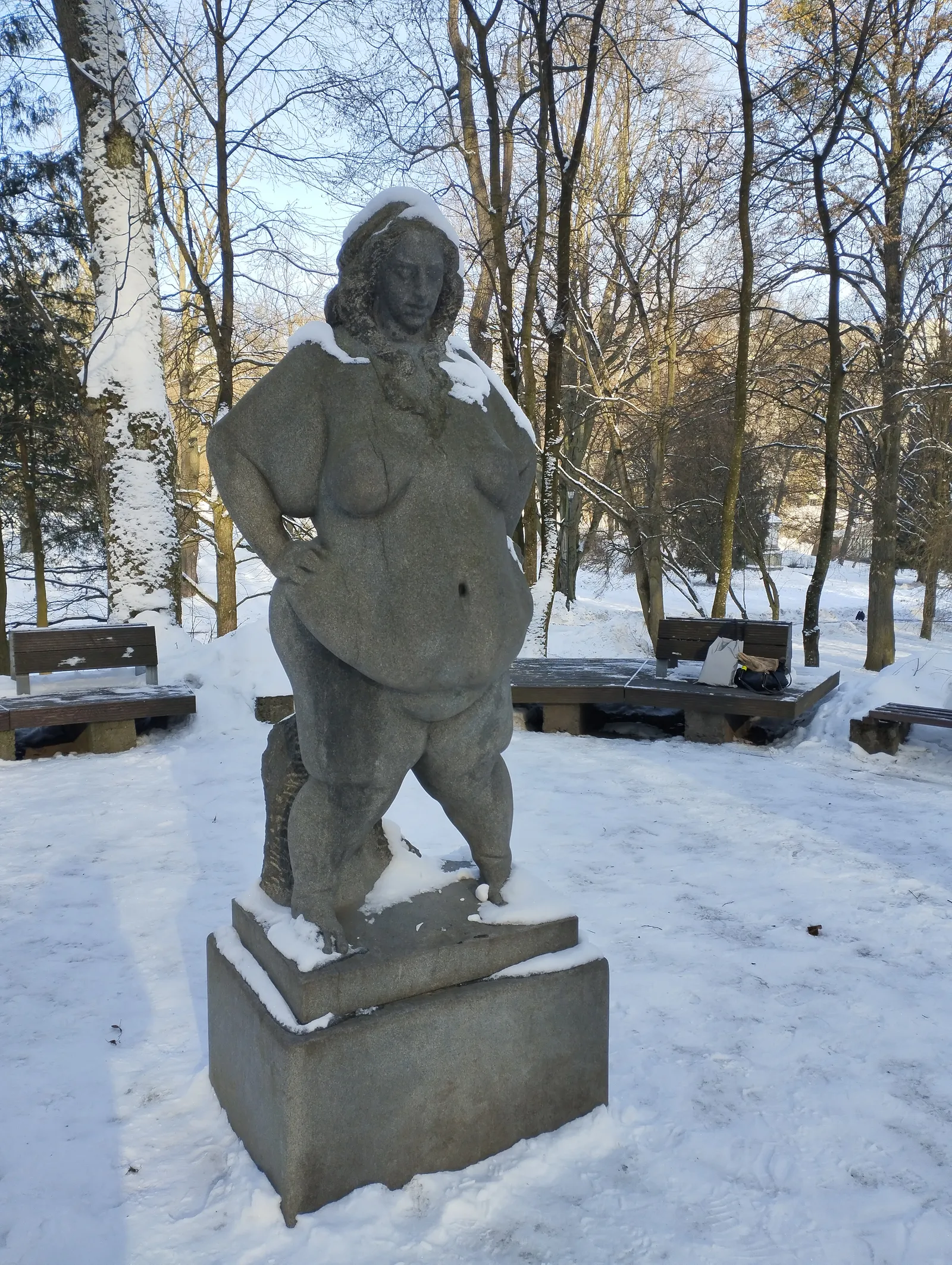 A frontal view of the granite statue 'Confident' standing on its stone pedestal in a snowy park: a nude, full-figured woman with one hand resting on her hip and snow settled across her shoulders and hair, park benches and bare trees in the background.