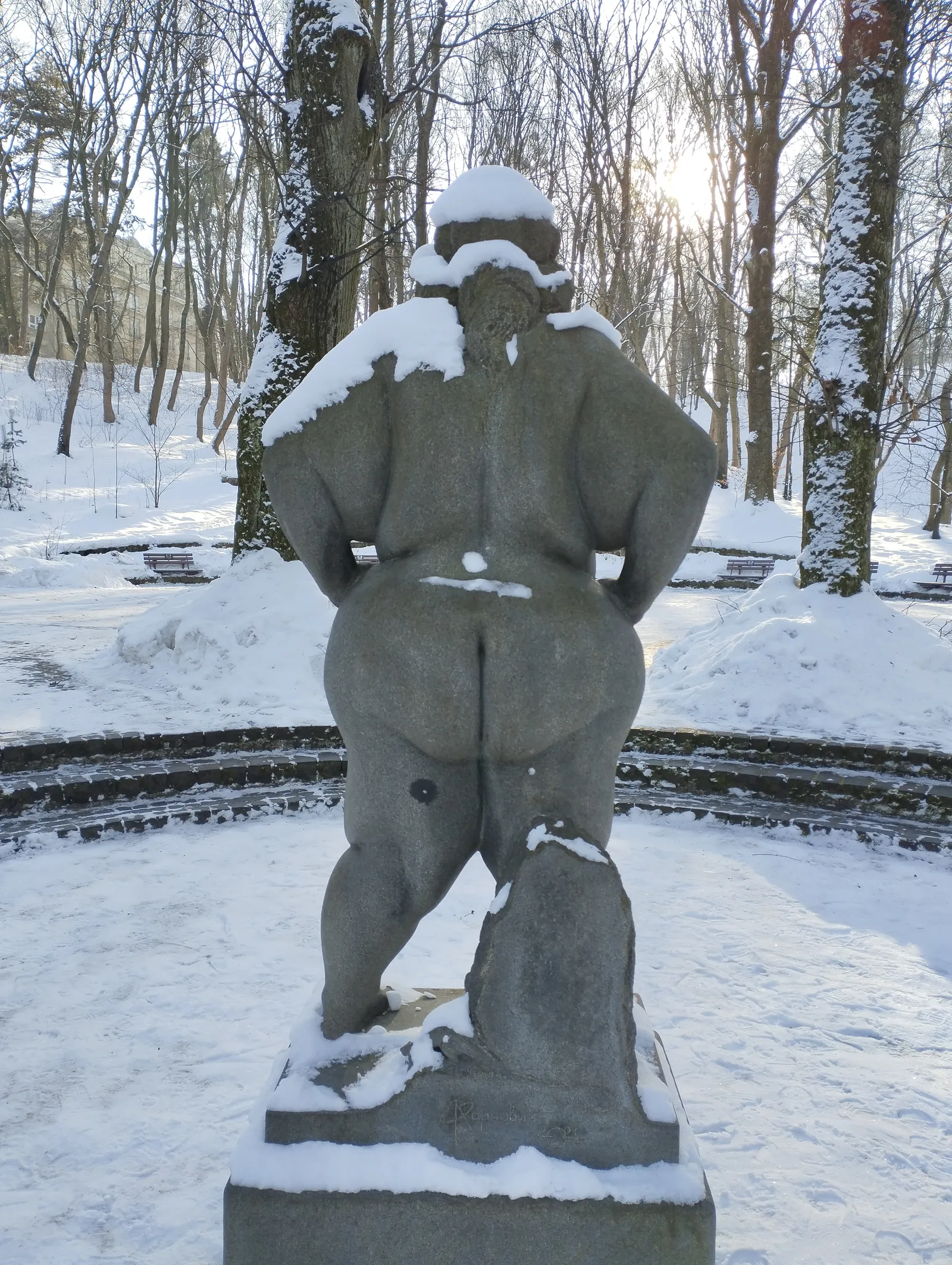 A rear view of the granite statue 'Confident' on its snow-covered pedestal in Stryi Park, Lviv: seen from behind, a nude, full-figured woman with snow settled across her shoulders and in her carved hair, both hands resting near her hips, the sculptor's signature and a date carved into the stone base below her.