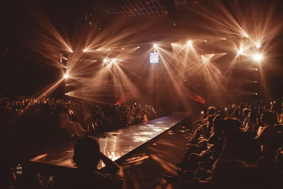 A packed runway show at night, spotlights sweeping over the crowd on both sides of a long catwalk.