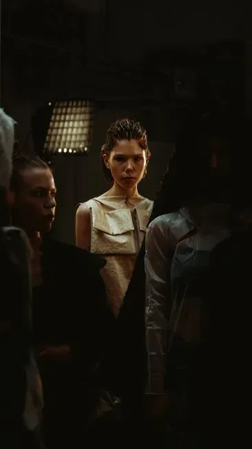 Two models waiting backstage in low light before a show, one in a pale structured top looking past the camera.