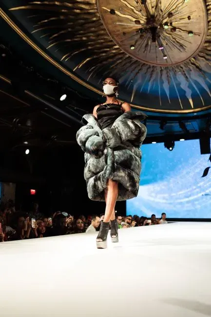 A model in an oversized grey fur coat and platform heels walking a runway under an ornate gold ceiling installation.