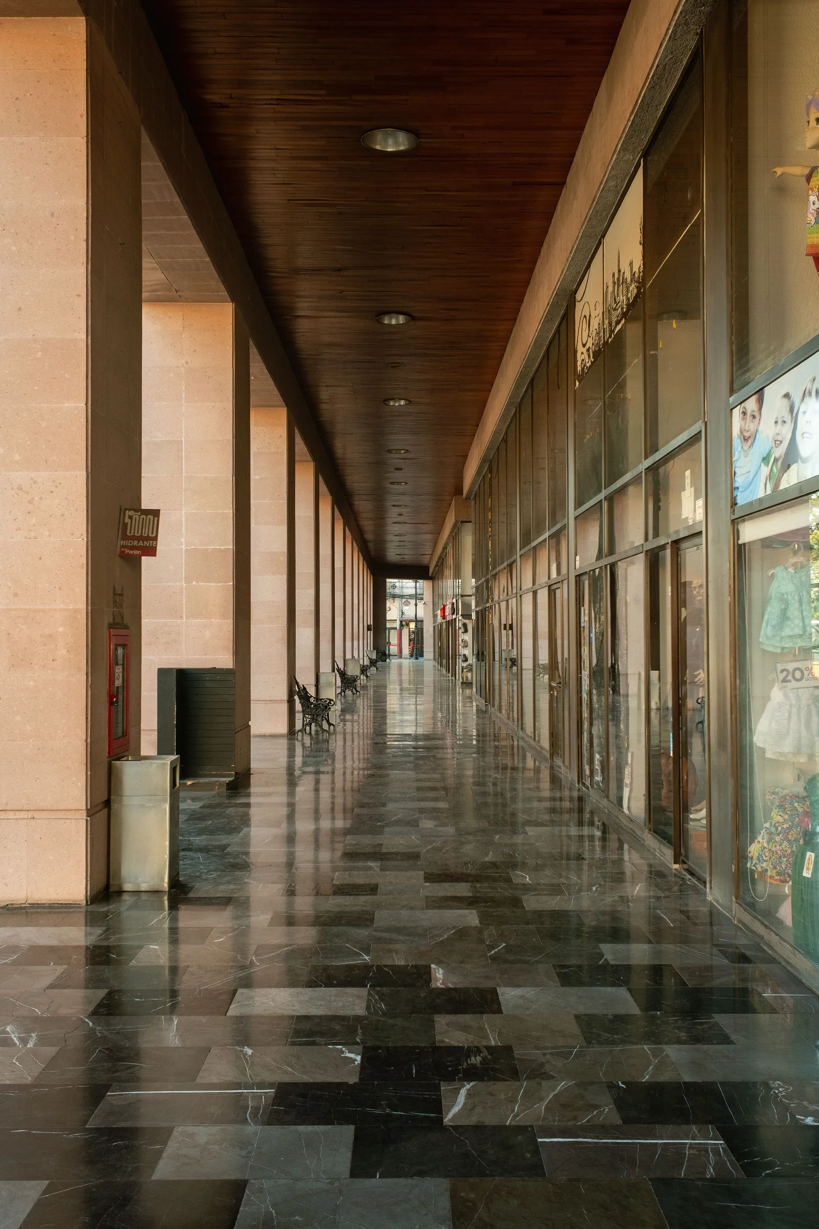 A long, empty shopping arcade with a polished chequered stone floor, lit shop windows down one side and no shoppers in sight.